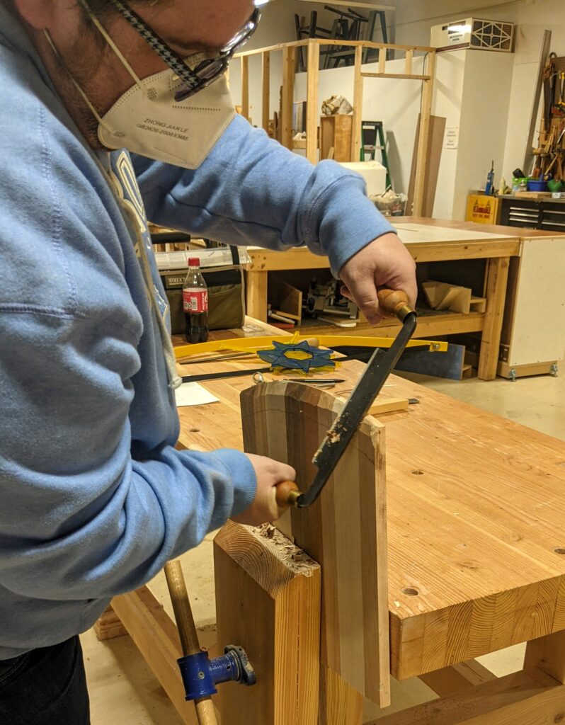A person in a blue hoodie uses a draw knife to remove wood for the bevel.