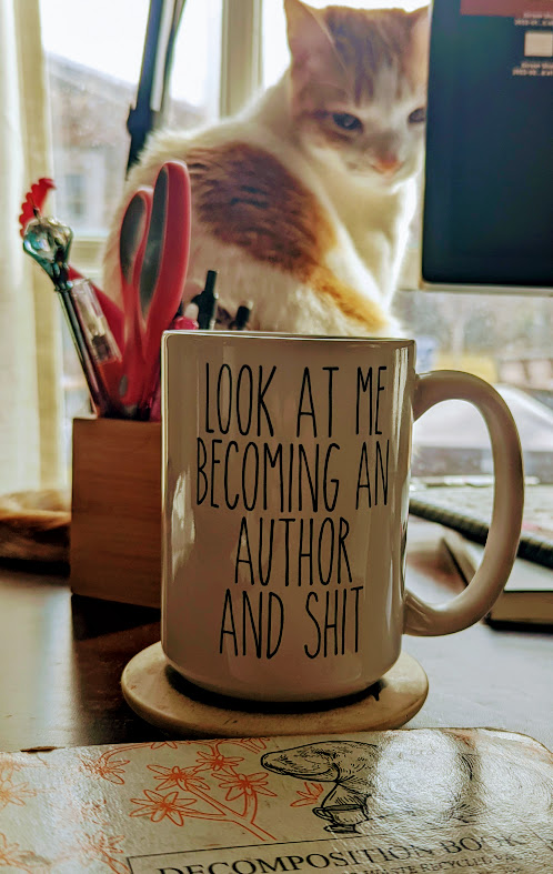 A cat looking at the camera, with a mug that says "Look at me becoming an author and shit" and a notebook.