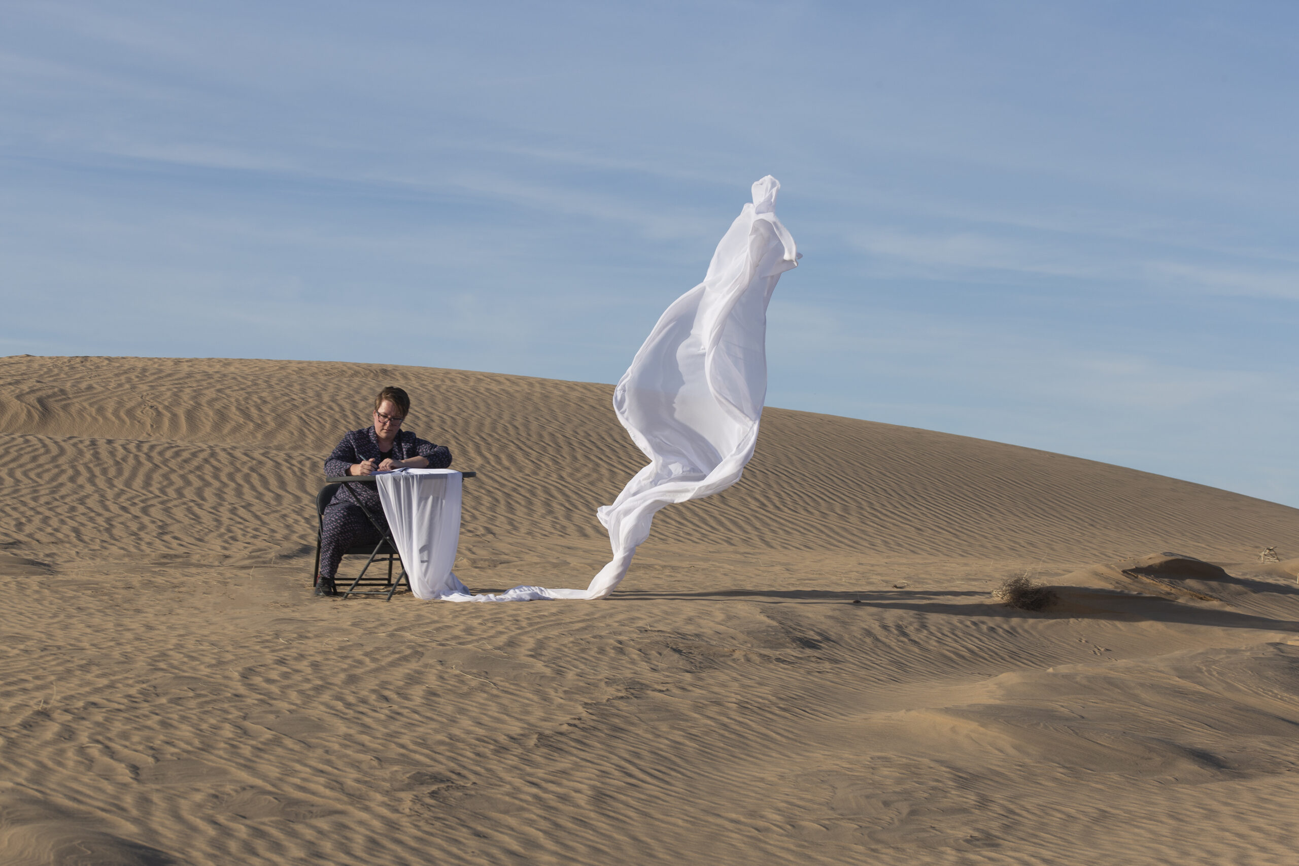 Tara sits on a sand dune, writing by hand. A piece of white fabric flows away from the paper and flies in the wind. Tara is wearing a patterned suit.